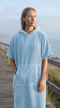 Woman wearing a light blue hooded poncho on a wooden boardwalk by the beach.
